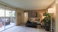 This single-floor space centers around a well-appointed living room that balances comfort and functionality. The room features neutral beige carpeting and walls complemented by a warm taupe accent wall, creating a layered, inviting atmosphere. Natural light flows generously into the space through a large sliding glass door fitted with sheer floor-to-ceiling drapes, which opens to an outdoor balcony or patio area, offering a seamless connection to the outdoors. A wall-mounted air conditioning unit ensures year-round climate comfort. The living area is thoughtfully arranged with a classic white sofa accented by decorative pillows, anchored by a dark wood coffee table that adds contrast to the light flooring. A vibrant abstract artwork on the accent wall serves as a focal point, adding a splash of color to the otherwise neutral palette. An additional window in the adjacent hallway or room brings in supplementary natural light, enhancing the overall brightness of the space. Lush indoor greenery adds a refreshing, organic touch to the interior. The layout appears practical and open, offering easy flow between the living area and the rest of the home.