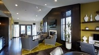 This single-floor space showcases a sleek, contemporary design with a striking contrast between dark and light elements. The main lounge area is anchored by a modern fireplace set against a rich, dark wood-paneled accent wall, creating a bold focal point. A wall-mounted flat-screen television sits above the fireplace, adding to the room's modern aesthetic. Seating is arranged thoughtfully around a low-profile round coffee table, featuring plush gray upholstered armchairs and sofas that invite comfortable gathering. A vibrant chartreuse area rug adds a bold pop of color against the dark hardwood flooring, energizing the space with a contemporary flair. Tall plantation shutters on the floor-to-ceiling windows allow for flexible light control while maintaining privacy, and a set of French doors opens to an outdoor area, flooding the space with natural light. Recessed lighting in the vaulted white ceiling provides warm, ambient illumination throughout. A statement indoor plant in a white pot brings a natural, organic touch to the otherwise structured interior. In the background, a dining or bar area is visible, reinforcing the space's versatility and open, flowing layout.