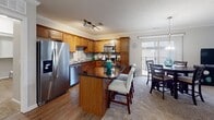 This single-floor residence is thoughtfully designed with an open-concept layout that seamlessly connects the kitchen, dining area, and living room. The kitchen is outfitted with warm honey-toned cabinetry, stainless steel appliances, a dark granite-topped center island with bar seating, hardwood flooring, and a tile backsplash. Crown molding and track lighting add refined detail overhead. The dining area sits adjacent to the kitchen, benefiting from natural light streaming through a glass door leading to a balcony. The living room features plush carpeting, a textured ceiling, crown molding, a ceiling fan, and a striking teal accent wall that anchors the space with color and character. Multiple bedrooms are carpeted and generously sized, each equipped with a ceiling fan and natural light from window treatments. Closet access is present in the secondary bedroom. The two full bathrooms share a consistent design language, featuring rich wood vanities with dark granite countertops, framed mirrors, and wood-look flooring. One bathroom includes a tub-shower combination. Together, the rooms offer a cohesive, comfortable living environment with consistent finishes throughout.