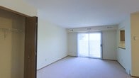 This single-floor space features a clean, open living area finished with wall-to-wall carpeting in a neutral tone that complements the light-colored walls throughout. The room is anchored by a sliding glass door fitted with vertical blinds, which serves as the primary source of natural light, brightening the interior and offering direct outdoor access. The ceiling is smooth and finished with a subtle texture, maintaining a clean, uniform appearance. HVAC vents are integrated into both the ceiling and walls, ensuring efficient climate control throughout the space. Trim detailing along the base of the walls adds a polished finish to the room. A wood-toned interior door is visible to one side, suggesting access to an adjacent room or closet. The overall layout is straightforward and versatile, offering an unobstructed floor plan well-suited to a variety of furniture arrangements. The neutral color palette and soft carpeting contribute to a calm, understated ambiance throughout the space.