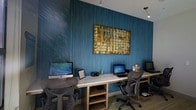 This single-floor business center is designed to provide a functional and visually engaging workspace. The room is anchored by a long, light wood workstation desk that spans the feature wall, offering multiple computer setups — including an Apple iMac and additional desktop computers — making it well-equipped to accommodate several users simultaneously. Ergonomic mesh chairs at each station ensure comfort during extended work sessions. The standout design element is the deep teal textured accent wall, which lends a bold and contemporary atmosphere to the space. Mounted prominently above the workstations is a large, colorful abstract painting with warm golden and earthy tones, providing a striking visual contrast against the cool blue backdrop. Track lighting suspended from the ceiling illuminates the workspace efficiently, highlighting both the artwork and the desks below. The flooring features a patterned carpet that complements the overall color palette. Integrated shelving beneath the main desk provides additional storage for equipment and supplies. Overall, this business center strikes a balance between professional utility and polished interior design, creating an inviting environment suited for productivity.