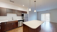 This thoughtfully designed 2-bedroom, 2-bathroom single-floor residence offers a functional and comfortable layout across a generous footprint. The open-concept kitchen is a standout feature, showcasing rich dark-wood cabinetry, a full suite of stainless steel appliances including a range, microwave, dishwasher, and refrigerator, and a central island with a light-toned countertop illuminated by stylish pendant lighting. Wood-look flooring flows seamlessly from the kitchen into the living room, which is bright and airy, benefiting from natural light through sliding glass doors that open to an outdoor area. The primary bedroom is spaciously proportioned, featuring plush carpeting, a window with natural light, and direct access to a private bathroom. The second bedroom, also carpeted, offers a comfortable and versatile space. Both bathrooms are finished with matching dark-wood vanities, white countertops, large mirrors with overhead lighting, and wood-look tile flooring. One bathroom includes a walk-in shower. A dedicated closet and hallway complete the layout, providing practical storage and connectivity throughout the residence. The overall design maintains a clean, neutral palette that is both modern and welcoming.
