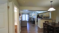 Located at 650 Bentley Ridge Boulevard in Lancaster, Pennsylvania, this single-floor home offers a well-designed, open-concept layout that seamlessly connects the main living areas. Rich, dark hardwood flooring runs throughout the space, providing a warm and cohesive foundation for the interior. The living room is bright and welcoming, featuring large windows with floor-length curtains that allow ample natural light to fill the room. A sliding glass door enhances the sense of openness and provides access to the outdoors. The kitchen is positioned adjacent to the living area, separated by a breakfast bar with stone countertops that doubles as a casual dining space. Dark cabinetry lines the kitchen walls, complemented by stainless steel appliances including a refrigerator, microwave, and additional built-in fixtures. A dedicated dining area with seating is conveniently situated near the kitchen, ideal for everyday meals or entertaining. Structural columns add an architectural detail to the open floor plan without disrupting the flow between spaces. A hallway leads to additional rooms, including a bathroom visible from the main corridor, fitted with classic fixtures and warm wood-toned cabinetry. Overall, the home presents a functional and comfortable living environment with a clean, neutral palette throughout.