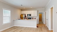 This thoughtfully designed single-floor home offers 2 bedrooms and 2 bathrooms within a comfortable and functional layout. The open-concept kitchen and living area serves as the heart of the home, featuring light hardwood flooring, crown molding, and recessed lighting throughout. The kitchen is equipped with stainless steel appliances — including a refrigerator, range, and microwave — complemented by crisp white shaker cabinetry, light stone countertops, and a peninsula with a built-in sink that provides additional prep space and seating. The living area receives ample natural light through multiple windows fitted with white shutters. Both bedrooms feature soft carpeting, crown molding, ceiling fans, and natural light from windows with blinds. The primary bedroom is generously sized and connects to its own bathroom. The second bathroom includes a single-sink vanity with a white cabinet, a mirror, and a tub-shower combo. A dedicated laundry room houses a full-size washer and dryer with overhead wire shelving. Consistent use of light gray walls, white trim, and light hardwood flooring throughout creates a cohesive, clean aesthetic across the entire home.