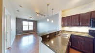 This single-floor space showcases an open-concept layout that seamlessly connects the kitchen and living area, creating a cohesive and functional environment. The kitchen is appointed with rich, dark-stained cabinetry that provides ample storage, complemented by sleek granite countertops in a deep, speckled finish. A double-basin sink sits along the countertop, and a peninsula-style breakfast bar extends into the living space, making it ideal for casual dining or entertaining. Pendant lighting hangs above the peninsula, adding a modern decorative touch, while a track lighting fixture further illuminates the kitchen zone. The flooring throughout features hardwood-style planks that add warmth and continuity across the open space. The living area benefits from a large window that draws in natural light and offers views of the surrounding exterior. Contrasting wall colors, including a cool grey accent wall, add visual depth and a contemporary feel to the overall aesthetic. The layout maximizes the sense of space while maintaining a well-defined flow between the kitchen and living areas.