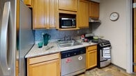 Located at 108 Dryden Rd in Ithaca, New York, this single-floor residential unit offers a well-organized layout designed to accommodate multiple occupants comfortably. The space features several private bedrooms, each carpeted and equipped with a bed, dresser, and a window fitted with horizontal blinds. Many of the bedrooms enjoy elevated views of the surrounding Ithaca landscape, with lush greenery and the city visible in the distance — a notable highlight of the unit. The living area provides a cozy setting with a sofa, coffee table, and a large window that further frames the scenic outdoor views. Adjacent to the living space, the dining area includes a wooden table and chairs alongside a wall-mounted television, creating a practical shared gathering space. The kitchen is outfitted with warm wood cabinetry, granite-style countertops, a stone tile backsplash, a stainless steel dishwasher, a microwave, and a range — offering ample functionality for everyday meal preparation. The full bathroom includes a shower-tub combination, a vanity with under-sink storage, a toilet, and mirrored medicine cabinets. Carpeted floors run throughout the bedrooms and common areas, contributing to a comfortable and cohesive atmosphere throughout the unit.
