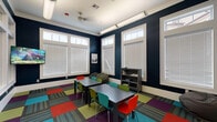 This energetic single-floor space is designed to inspire creativity and collaboration. The room is defined by striking navy blue walls accented with crisp white trim and crown molding, creating a bold yet polished aesthetic. Large windows fitted with white horizontal blinds line multiple walls, allowing generous amounts of natural light to fill the interior throughout the day. Transom windows above the main panes further enhance the brightness of the space.

The flooring is a standout feature, composed of vibrant modular carpet tiles in a patchwork arrangement of red, teal, green, purple, and striped patterns, adding a dynamic and playful energy underfoot. Two rectangular gray tables serve as the central workspace, surrounded by a mix of colorful chairs in red, green, and teal, providing comfortable seating for group activities or study sessions.

A gray bookcase along one wall holds books and supplies, while a wall-mounted flat-screen television offers multimedia capabilities. Bean bag chairs tucked into corners provide casual, flexible seating options. Overhead, elongated tubular light fixtures supplement the natural light. Large interior windows on one side offer sightlines into an adjacent fitness area, reinforcing the sense of openness. The overall ambiance is lively, functional, and welcoming.