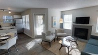 This single-floor space offers a welcoming and well-appointed interior designed for comfortable everyday living. The living area serves as the heart of the home, bathed in natural light streaming through multiple windows dressed with classic white horizontal blinds. The warm hardwood-style flooring extends throughout, adding a sense of warmth and continuity to the space.

The living room is thoughtfully arranged with a pair of stylish patterned accent chairs set atop a woven area rug, centered around a sleek glass-top coffee table — creating an inviting conversational setting. A floor lamp provides additional ambient lighting, complementing the natural brightness of the room. A framed abstract artwork adorns the wall, adding a touch of contemporary style.

A notable feature of the living area is the classic fireplace framed by a traditional mantel, offering both a functional and aesthetic focal point. An exterior door with glass panels leads to the outside, further enhancing the flow of natural light. The neutral gray walls and textured ceiling tie the space together with a clean, modern aesthetic. Overall, the space strikes a balance between casual comfort and refined style, making it a versatile and functional home.