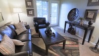 This single-floor space centers around a well-appointed living area that balances comfort and style. The room features a dark leather sofa and matching armchair arranged around a dark wood coffee table, creating an inviting seating arrangement suited for relaxation or conversation. A richly patterned area rug in warm earth tones anchors the space and adds a bold visual element beneath the furniture grouping. Natural light flows generously into the room through multiple large windows fitted with horizontal blinds, as well as a glass-paneled door that offers a view of lush greenery outside, enhancing the connection between interior and exterior. The neutral-toned walls provide a clean backdrop that allows the furnishings and décor to stand out. A dark console table positioned along one wall displays a striking circular decorative piece alongside dark ceramic vases, contributing an artistic focal point. Framed artwork adorns the walls, adding a polished, curated feel to the space. The carpeted flooring extends throughout, contributing to the overall warmth of the room. Together, these elements create a cohesive, intimate living environment with thoughtful attention to design detail.