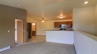 This single-floor residence offers a thoughtfully designed open-concept layout that seamlessly connects the living and kitchen areas. The living space features soft, wall-to-wall carpeting that adds warmth and comfort underfoot, while a ceiling fan overhead provides both practical airflow and a subtle decorative touch. A smooth transition between carpet and tile flooring marks the boundary between the living area and the kitchen zone. The kitchen is equipped with warm, medium-toned wood cabinetry that provides generous storage, complemented by stainless steel appliances including a microwave and range. A peninsula counter with a stone-look surface extends into the open space, offering a casual dining or prep area while maintaining visual connectivity throughout the layout. The neutral, light-toned walls and ceiling throughout the space contribute to a clean, airy ambiance. A corridor visible from the main living area leads to additional rooms, including a bathroom that features its own vanity and lighting. The overall design is functional and cohesive, with consistent finishes that create a unified, comfortable living environment across the entire floor.