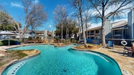 Pool 2 at 2700 Central Drive in Bedford, Texas is a well-appointed outdoor aquatic amenity space designed for relaxation and recreation. The centerpiece is a generously sized freeform swimming pool with vivid turquoise water, shaped with sweeping curves that create a resort-style aesthetic. Natural stone rock formations are incorporated throughout the pool's perimeter, adding a distinctive, naturalistic character to the design. A gradual wading entry with traction-grip steps allows for easy and safe access into the pool, while a separate circular spa area with stone coping offers a dedicated space for relaxation. The pool deck is finished with stone and concrete pavers, providing ample space for lounge chairs and seating arrangements positioned to take full advantage of the open sky and abundant natural sunlight. Shade umbrellas and additional seating areas are positioned along the perimeter, offering comfortable spots to unwind. The area is enclosed by a wrought iron security fence, ensuring safety while preserving open sightlines to the surrounding mature trees and residential buildings. Lush landscaping, including ornamental grasses and native plantings, frames the space throughout. The overall ambiance is that of a relaxed, resort-inspired outdoor retreat set within a well-established community in Bedford, Texas.