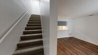 Located in Austin, TX, this two-story, 1-bedroom, 1-bathroom unit offers a practical and well-organized layout spread across two floors. The main level features a living room with warm wood-look flooring, clean white walls, a textured ceiling, and a sliding glass door that fills the space with natural light and provides views of the surrounding greenery. Adjacent to the living area is a galley-style kitchen equipped with gray shaker cabinets, a stainless steel refrigerator, a black electric range, and a double-basin sink set against light countertops — offering ample storage and functionality in a compact footprint. A carpeted staircase connects the two floors, leading up to the second level where the generously sized bedroom awaits. The upper floor features the same wood-look flooring, bright white walls, a ceiling fan, and a closet with open shelving. The full bathroom is located on this level and includes a tiled shower/tub combo, a toilet, and a gray vanity cabinet for additional storage. Throughout the unit, the textured ceilings, neutral color palette, and consistent flooring create a cohesive and practical living environment.