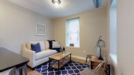 This single-floor unit offers a thoughtfully designed layout that combines industrial and farmhouse-inspired aesthetics across its key living areas. The kitchen is a functional centerpiece, featuring white cabinetry, dark granite countertops, stainless steel appliances including a range, microwave, refrigerator, and dishwasher, along with warm wood-tone luxury vinyl plank flooring and open floating shelves with pendant lighting overhead. A galley-style configuration maximizes efficiency while maintaining an open, airy feel. The living area provides a comfortable retreat, furnished with a light-toned sofa accented by navy blue throw pillows, a rustic wood-and-metal coffee table, and a bold navy geometric-patterned area rug. A window allows natural light to fill the room, complemented by a cage-style ceiling fixture and exposed ductwork that add an industrial touch. The bedroom is a serene space with plush wall-to-wall carpeting, a classic black iron bed frame, a whitewashed dresser, and decorative wall art that reinforces the farmhouse character. The bathroom is clean and modern, showcasing a dark espresso vanity with a white countertop, a framed mirror, black hardware accents, wood-look flooring, and a glass-enclosed shower, creating a polished, cohesive finish throughout.