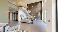 Located at 1515 Rollick Rd in Cary, NC, this two-story facility spans a generously sized footprint across two levels, with the main floor offering the majority of the space. The ground floor makes an immediate impression with a dramatic double-height lobby featuring floor-to-ceiling black-framed windows that flood the interior with natural light and offer views of the surrounding wooded landscape. A sweeping staircase with glass railings connects the two levels, anchored by a striking wood-paneled accent wall and an eye-catching sculptural chandelier. The lounge area is furnished with plush seating, patterned area rugs, and curated artwork, creating an inviting and upscale ambiance. Steel-framed glass partition walls define enclosed conference and meeting rooms, each equipped with modern pendant lighting, dark conference tables, and wall-mounted displays. A catering or refreshment area is accessible from the conference space. The private office features a granite-topped desk, designer carpet, and a linear pendant fixture. Hallways are finished with large-format tile flooring and gallery-style wall art, including a distinctive arrangement of local area maps. The upper level provides additional hallway and support space, overlooking the grand lobby below. Throughout, the design balances contemporary architecture with refined, curated interiors.