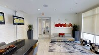 This single-floor space presents a sleek, contemporary lobby or reception area designed with a clean, modern aesthetic. The interior is characterized by bright white walls and light-toned tile flooring that together create an open, airy atmosphere. Recessed lighting is embedded throughout the ceiling, providing consistent, well-distributed illumination across the space.

A central seating area anchors the room, featuring a light gray sofa accented with decorative throw pillows in red and patterned tones. Two natural stone-topped side tables rest atop a stylized area rug with an abstract gray and white pattern, adding texture and visual interest to the floor. Bold abstract artwork in red and orange hues adorns the wall above the seating area, serving as a striking focal point.

Tall, dark-toned planters housing upright tropical plants are positioned symmetrically throughout the space, introducing natural elements and enhancing the room's contemporary character. A reception or check-in desk is partially visible in the foreground, and a glass-paneled door leads to an adjacent interior space. A wall-mounted screen is also present, suggesting a functional, multi-purpose use for this welcoming area.