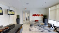 This single-floor space presents a sleek, contemporary lobby or reception area designed with a clean, modern aesthetic. The interior is characterized by bright white walls and light-toned tile flooring that together create an open, airy atmosphere. Recessed lighting is embedded throughout the ceiling, providing consistent, well-distributed illumination across the space.

A central seating area anchors the room, featuring a light gray sofa accented with decorative throw pillows in red and patterned tones. Two natural stone-topped side tables rest atop a stylized area rug with an abstract gray and white pattern, adding texture and visual interest to the floor. Bold abstract artwork in red and orange hues adorns the wall above the seating area, serving as a striking focal point.

Tall, dark-toned planters housing upright tropical plants are positioned symmetrically throughout the space, introducing natural elements and enhancing the room's contemporary character. A reception or check-in desk is partially visible in the foreground, and a glass-paneled door leads to an adjacent interior space. A wall-mounted screen is also present, suggesting a functional, multi-purpose use for this welcoming area.