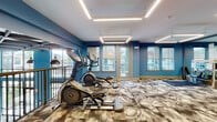 Located at 7200 N Frontage Rd in Orlando, FL, this two-floor fitness center is designed to support a comprehensive workout experience. The space is defined by its striking blue accent walls, patterned carpet flooring, and modern suspended linear lighting that illuminates every corner with a clean, energetic ambiance.

The ground floor features soaring double-height ceilings with expansive floor-to-ceiling windows and glass doors that flood the space with natural light and offer views of the surrounding outdoor landscape. This level is equipped with a wide variety of fitness equipment, including rowing machines, a StairMaster, battle ropes, cable machines, weight benches, and a dedicated free weights and dumbbell rack area. Structural columns are wrapped in blue, reinforcing the facility's vibrant design language.

The upper floor offers additional workout zones, including elliptical machines, a Pilates/reformer area with a HyperIce recovery station, stretching mats, and full-length mirrors that visually expand the space. Geometric pendant lighting and glass-paneled French doors connect the areas seamlessly. An elevator provides accessible vertical circulation between floors.

Together, the two levels provide a well-rounded, thoughtfully equipped fitness facility with a modern aesthetic tailored for a diverse range of training modalities.