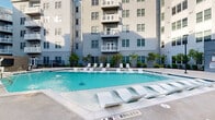 Located at 3061 George Busbee Pkwy NW in Kennesaw, Georgia, this outdoor amenity space is designed to deliver a resort-style living experience within a contemporary multi-story residential community. At the heart of the space is a generously sized swimming pool with crystal-clear turquoise water, framed by a wide, well-maintained concrete pool deck. The pool features a shallow sun shelf fitted with sleek, curved white lounge chairs, allowing residents to relax partially in the water. Additional white chaise lounges are arranged along the perimeter of the deck, providing ample space for sunbathing. The courtyard is enclosed by the multi-story building on multiple sides, creating a sense of privacy and intimacy while offering views of resident balconies above. A shaded gathering area with a blue canopy sail and an adjacent clubhouse entrance, marked with bold blue architectural accents and string lighting, adds a social focal point to the space. Thoughtfully placed landscaping — including ornamental trees, sculpted shrubs, and mulched planting beds — lines the building facade, softening the hardscape and adding a touch of greenery throughout. Iron fencing defines the pool zone, while classic streetlamp-style lighting posts complete the polished, community-centered aesthetic of this outdoor retreat.