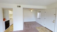 This single-story space presents a straightforward, functional layout with a clear distinction between living areas. The main living area transitions from tile flooring near the entryway into wall-to-wall carpeting that extends throughout the adjoining room, creating a comfortable and cohesive feel underfoot. White walls throughout the space lend a clean, neutral backdrop that allows for versatile decorating possibilities.

A textured popcorn ceiling adds a classic architectural detail overhead, while a ceiling fan with a built-in light fixture is centrally positioned in the carpeted room, providing both air circulation and ambient lighting. A window fitted with horizontal blinds allows natural light to filter into the space, brightening the interior while maintaining privacy.

The open passage between the tiled and carpeted areas creates a sense of connected, flowing space across the floor plan. HVAC vents are integrated into both the ceiling and walls, ensuring efficient climate control throughout. The overall ambiance is simple and move-in ready, offering a blank canvas well-suited for personalization.