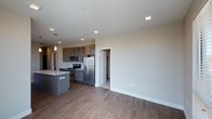 This well-appointed single-floor residence showcases a thoughtfully designed open-concept layout that seamlessly blends the kitchen and living areas into one cohesive, light-filled space. The kitchen is a standout feature, outfitted with a full suite of stainless steel appliances including a refrigerator, range, and microwave. Dark, rich cabinetry provides ample storage and contrasts beautifully against the light countertops and neutral wall tones. A kitchen island with a built-in sink adds both functionality and a natural gathering point, accented by a pendant light that contributes a warm, modern touch. Recessed lighting throughout the ceiling ensures consistent, even illumination across the open floor plan. Durable hardwood-style flooring in a warm medium-tone finish extends through the kitchen and living areas, adding texture and visual warmth to the space. The living area offers generous room for furnishing, with clean white walls and large baseboards lending a crisp, polished feel. A doorway leads to an additional room, suggesting further private living or sleeping quarters. The overall ambiance is modern, clean, and move-in ready, ideal for those who appreciate contemporary design and functional living spaces.