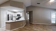 This contemporary residence showcases an open floor plan with vaulted ceilings, creating a spacious atmosphere. The kitchen boasts white cabinetry, stainless steel appliances, and a herringbone backsplash. Wood-look flooring extends through main living areas, while bedrooms feature plush carpeting. Bathrooms are equipped with modern fixtures and tiled surfaces. Neutral tones dominate the interior, complemented by ample natural light from windows. Ceiling fans in bedrooms provide comfort. The layout includes multiple bedrooms, bathrooms, and living spaces, offering functionality and style in a single-story design.