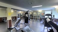 This fitness center is designed to accommodate a comprehensive strength and conditioning workout. The space is outfitted with a notable selection of equipment, including a multi-function cable machine, adjustable weight benches, a power rack with barbell and plates, and additional free weight stations. The rubber flooring provides a durable, slip-resistant surface suited for heavy lifting and high-intensity training. Large mirrors line the walls, visually expanding the space while allowing users to monitor their form during exercises. Overhead fluorescent lighting ensures the area is evenly and brightly illuminated throughout. The concrete block walls contribute to the utilitarian character of the gym, emphasizing its function-forward design. The layout is organized to allow users to move between stations with ease, making efficient use of the available floor space. Overall, the fitness center offers a practical and well-appointed environment suitable for a range of strength training routines.