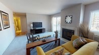 This single-floor residence is thoughtfully laid out to maximize functionality across its key living spaces. The living room serves as the central gathering area, finished with plush carpeting and neutral-toned walls. A fireplace with a clean surround anchors one wall, while sliding glass doors open to a private balcony, allowing natural light to fill the space and offering outdoor views. The room comfortably accommodates both seating and media areas.

Adjacent to the living room, the dining area features carpeted flooring and is illuminated by a pendant light fixture overhead. A pass-through opening connects directly to the kitchen, which is characterized by its bold black-and-white checkered tile floor, warm wood cabinetry, white appliances including a gas range, and ample counter space.

The primary bedroom is generously sized with carpeted floors, a window for natural light, and mirrored sliding closet doors that open to a well-organized walk-in storage area. The second bedroom is similarly carpeted and bright, featuring a window with blinds. Both bathrooms include pedestal sinks, tile flooring, and large vanity mirrors—one with a tub/shower combination and one as a half bath configuration.