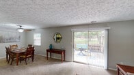 This single-story residence offers a thoughtfully laid-out floor plan with multiple living spaces, each finished with a textured ceiling and neutral gray walls throughout. The carpeted living room serves as the heart of the home, bathed in natural light from a large sliding glass door that opens to a patio area overlooking a green outdoor space — ideal for indoor-outdoor living. An adjacent dining area accommodates a full-sized table and is equipped with a ceiling fan and natural light from a side window. The kitchen features white cabinetry with wood trim detailing, white appliances including a range and over-the-range microwave, light countertops, and a double basin sink, all set on tile flooring. The primary bedroom includes a striking deep teal accent wall and plush carpet, while a second bedroom showcases an eye-catching full-wall floral mural, giving it a creative, versatile character suited as a bedroom or flex space. Two bathrooms, each with a vanity, large mirror, and Hollywood-style lighting, serve the bedrooms conveniently. The overall ambiance is comfortable and functional, with consistent neutral tones providing a cohesive feel throughout.