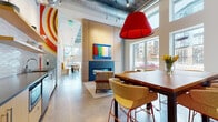 This single-floor amenity space makes a striking impression through its blend of industrial architecture and vivid, curated design. Polished concrete floors run throughout, unifying distinct functional zones beneath exposed ceilings with visible ductwork and concrete structural elements that lend an urban, loft-like character.

The main lounge area is a standout, bathed in natural light from expansive floor-to-ceiling windows that frame views of the surrounding cityscape. Bold red pendant lights anchor the space visually, complementing an eclectic mix of mid-century-inspired furnishings in warm yellows, rich leathers, and patterned textiles. A built-in fireplace with a teal surround, topped by a colorful abstract painting, adds a warm focal point, while a large communal dining table nearby provides a collaborative gathering spot.

The entry foyer features black steel-framed glass doors, a sculptural globe chandelier, and curated wall art, establishing an inviting, gallery-like arrival experience. A well-appointed kitchenette includes warm wood cabinetry, stone countertops, a decorative tile backsplash, and stainless steel appliances.

The fitness center is generously scaled, outfitted with treadmills, stair climbers, stationary bikes, free weights, and wall-mounted TVs — all set against mirrored walls and polished concrete floors.
