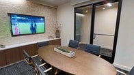 This polished single-floor conference room offers a refined and professional environment designed for productive meetings and collaboration. At the center of the space sits a generously sized oval walnut table, its warm wood tones complemented by an arrangement of sleek black leather chairs that provide comfortable seating for multiple attendees. A decorative live-moss centerpiece adds a subtle natural touch to the table surface.

The room's most striking architectural feature is a textured natural stone accent wall, which serves as a sophisticated backdrop for the wall-mounted flat-screen display — ideal for presentations, video conferencing, or media viewing. The neutral, light-toned walls contrast elegantly with the darker flooring, contributing to a clean and contemporary aesthetic.

A glass-paneled door with a dark metal frame connects the room to an adjacent corridor, maintaining visual openness while offering privacy when needed. Subtle recessed lighting illuminates the space efficiently, ensuring a well-lit yet comfortable atmosphere. A decorative vase with dried botanicals adds a finishing organic element, enhancing the room's overall warmth. Together, these design choices create a space that balances functionality with a modern, executive-level aesthetic.