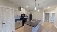 This single-floor residence located on Sherwood Avenue in Holland, Michigan, presents a clean, contemporary design throughout. The open-concept kitchen serves as a functional centerpiece, equipped with black appliances including a range, over-the-range microwave, and refrigerator. Crisp, light-toned cabinetry with matte black hardware contrasts against dark countertops, while a central island with a stainless steel sink provides additional workspace. Two pendant lights hang above the island, enhancing both practicality and visual appeal. Light wood-look flooring extends through the kitchen and living areas, contributing to a bright, cohesive aesthetic.

The living area is generously sized and features sliding glass doors that open to a private balcony, allowing ample natural light and outdoor views. The bedrooms feature neutral carpet, wide windows with blinds, and closets with built-in shelving. The unit includes two bathrooms—one with a tub-shower combination and light flooring, and another featuring a standalone walk-in shower with chrome fixtures. Neutral warm-toned walls throughout unify the space, creating a calm, move-in-ready environment.