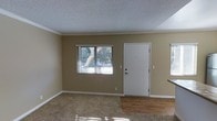 This single-story space presents a straightforward and functional layout highlighted by a combination of flooring materials. The main living area transitions between plush carpeting and hardwood-style flooring, offering both comfort and durability. Warm, neutral tan-colored walls create a understated backdrop throughout, complemented by crisp white baseboards and crown molding that add a touch of architectural detail. A textured popcorn ceiling runs overhead, a classic finish that adds subtle dimension to the space. The front entry features a solid white door flanked by a window dressed with horizontal blinds, allowing natural light to filter in while maintaining privacy. The window offers a glimpse of mature tree coverage outside, suggesting a pleasant outdoor setting. The entry area's hardwood-style flooring provides a clean and practical transition zone, while the carpeted living area adds warmth and softness underfoot. The overall ambiance is clean, neutral, and move-in ready, offering a blank canvas adaptable to a variety of interior styles and furnishing arrangements.