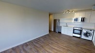 This single-floor space showcases a clean, modern design with an open-concept layout that seamlessly connects the living area to the kitchen. The flooring throughout is a warm-toned hardwood-style laminate, providing a cohesive and inviting aesthetic across the space.

The kitchen is positioned at the far end of the open layout and features white cabinetry with a minimalist design, offering ample storage. Stainless steel appliances, including a refrigerator, dishwasher, and stove, add a contemporary touch. A single-basin sink sits beneath a functional workspace, complemented by white subway tile backsplash that enhances the brightness of the area. Track lighting above the kitchen provides focused illumination over the workspace.

The living area is generously proportioned, with neutral grey walls that create a versatile backdrop suitable for a variety of interior styles. The space is well-lit and benefits from a clean, uncluttered design. A hallway visible in the background suggests additional rooms beyond the main living area. Overall, the space presents a practical and modern environment with a neutral palette.
