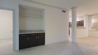 This single-floor space offers a well-organized and modern interior designed with both functionality and style in mind. The open layout creates a sense of spaciousness, with smooth white walls and light-toned carpeting that contribute to a bright and airy atmosphere throughout.

A notable feature is the built-in cabinetry unit set into a recessed alcove, showcasing rich dark wood-toned cabinets paired with a light stone or granite-style countertop. The contrast between the dark cabinetry and the light walls and flooring adds visual interest and a contemporary feel to the living area. An open shelf above the cabinet section provides additional display or storage options.

A structural column detail and open passageway connect the main living area to what appears to be a kitchen beyond, where additional dark cabinetry can be glimpsed. The ceiling features a recessed beam detail that adds architectural definition to the space, while ceiling vents indicate a central HVAC system for year-round comfort.

The overall ambiance of the space is clean, understated, and move-in ready, with a cohesive design language that runs throughout. The neutral tones and modern finishes make it a versatile canvas suited to a variety of interior styles and preferences.