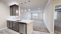 This well-appointed single-floor unit showcases a thoughtfully designed open-concept layout that seamlessly connects the kitchen and living area. The kitchen is a functional centerpiece, featuring rich dark-toned cabinetry, crisp white quartz countertops, a subway tile backsplash, and a full suite of stainless steel appliances including a gas range, microwave, refrigerator, and dishwasher. A peninsula with an undermount sink and elegant pendant lighting overhead enhances both the style and utility of the space. Light hardwood-style flooring runs through the kitchen and entry areas, transitioning to plush wall-to-wall carpeting in the living room and bedrooms, adding warmth and comfort underfoot. Crown molding and recessed lighting lend a refined architectural character throughout. The spacious living area is bathed in natural light through large windows, offering views of greenery outside. The bedrooms are generously proportioned, each with ample natural light from well-positioned windows. One bedroom features a door leading to an outdoor area. Neutral tones are carried consistently throughout the unit, creating a clean, cohesive, and move-in-ready atmosphere that is both modern and inviting.