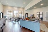 Located at 1633 Greenridge Dr in Burleson, TX, this single-floor community clubhouse is designed to impress with its open-concept layout and refined architectural details. Soaring vaulted and tray ceilings lend an airy, expansive feel throughout, while rich wood-look flooring grounds the space with warmth and continuity.

The main lounge area is a welcoming focal point, furnished with plush blue and gray sofas, leather accent chairs, and layered area rugs, all anchored beneath recessed lighting and a ceiling fan. A wall-mounted TV and an artfully crafted reclaimed wood media console add both function and character. Steel-framed glass partition doors offer a sophisticated separation between the lounge and an adjacent office or flex room.

Floor-to-ceiling steel-framed windows flood the space with abundant natural light and frame views of the surrounding community's stone-accented architecture and landscaping. A game area with a full-length shuffleboard table sits alongside these dramatic windows, creating an inviting recreational nook.

The kitchenette features granite countertops, dark cabinetry, stainless steel appliances, and a large island with bar seating — ideal for social gatherings. An eye-catching bicycle wheel wall installation adds a unique artistic touch. The dining area accommodates a generous table and chairs, completing this versatile and thoughtfully designed community space.