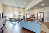 Located at 1633 Greenridge Dr in Burleson, TX, this single-floor community clubhouse is designed to impress with its open-concept layout and refined architectural details. Soaring vaulted and tray ceilings lend an airy, expansive feel throughout, while rich wood-look flooring grounds the space with warmth and continuity.

The main lounge area is a welcoming focal point, furnished with plush blue and gray sofas, leather accent chairs, and layered area rugs, all anchored beneath recessed lighting and a ceiling fan. A wall-mounted TV and an artfully crafted reclaimed wood media console add both function and character. Steel-framed glass partition doors offer a sophisticated separation between the lounge and an adjacent office or flex room.

Floor-to-ceiling steel-framed windows flood the space with abundant natural light and frame views of the surrounding community's stone-accented architecture and landscaping. A game area with a full-length shuffleboard table sits alongside these dramatic windows, creating an inviting recreational nook.

The kitchenette features granite countertops, dark cabinetry, stainless steel appliances, and a large island with bar seating — ideal for social gatherings. An eye-catching bicycle wheel wall installation adds a unique artistic touch. The dining area accommodates a generous table and chairs, completing this versatile and thoughtfully designed community space.