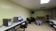 This single-floor space is designed to serve dual purposes, seamlessly combining a functional workstation area with a comfortable lounge. The room features neutral sage-green walls paired with light ceramic tile flooring, creating a clean and practical aesthetic throughout.

On one side, a long granite-topped desk runs along the wall, accommodating a desktop computer setup complete with a monitor, keyboard, and supporting equipment. A black leather office chair provides ergonomic seating at the workstation, making it well-suited for administrative or business tasks.

The lounge area on the opposite side is furnished with two black leather club chairs, a cushioned floral ottoman, and a brown sofa accented with decorative pillows. A wall-mounted flat-screen television is positioned to be easily visible from the seating area, enhancing the space's entertainment functionality.

The drop ceiling features recessed panels and a skylight that allows natural light to filter into the room, brightening the overall atmosphere. The layout is open and uncluttered, offering a versatile environment that can accommodate both productive work and relaxed social interaction.