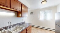 Located at 1401 E Bellows St in Mount Pleasant, Michigan, this single-floor unit offers a practical and well-organized layout. The galley-style kitchen is a standout feature, equipped with rich warm-toned wood cabinetry, dark granite-style countertops, a double stainless steel sink, and black appliances including a refrigerator, electric range, and dishwasher. Tile flooring runs throughout the kitchen and dining area, lending durability and easy maintenance to the high-use spaces. The kitchen opens toward a dining area brightened by a window with vertical blinds and a wall-mounted air conditioning unit, ensuring year-round comfort. Baseboard heating is present throughout the unit, providing consistent warmth during Michigan winters. The bedrooms feature soft carpeting, neutral wall tones, and windows that allow natural light to filter in. One bedroom includes a spacious closet with bi-fold doors and built-in hanging rods, offering ample storage. The overall layout, as seen in the dollhouse view, is elongated and efficiently designed, with clearly defined living, kitchen, and sleeping areas. The light, neutral color palette throughout creates a clean and versatile atmosphere, making the space easy to personalize.