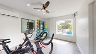 Located at 1250 Woodcrest Dr in Daytona Beach, FL, this dedicated exercise room offers a well-equipped fitness space designed for efficient, focused workouts. The room features light gray wood-look flooring and crisp white walls that create a clean, energizing atmosphere. A ceiling fan with integrated lighting provides overhead air circulation, complemented by a wall-mounted mini-split air conditioning unit to ensure comfortable workouts year-round. Recessed lighting adds to the bright, welcoming ambiance throughout the space. The room is outfitted with multiple SPT spin bikes positioned for cardio training, alongside a Smart Fitness multi-functional equipment rack stocked with a colorful assortment of medicine balls, stability balls, resistance bands, foam rollers, and additional accessories. Two windows allow natural light to fill the space — one offering a view of lush tropical greenery, and a larger picture window framing a poolside patio area with lounge seating, connecting the interior to the outdoor environment. A glass-panel door provides easy access to the exterior. The thoughtful layout maximizes the available footprint, making this a practical and inviting fitness facility suited for cardio and functional training.