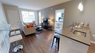 This single-floor residence showcases a thoughtfully designed open-concept layout that seamlessly connects the kitchen, dining, and living areas. The kitchen is outfitted with white shaker cabinetry, dark quartz countertops, a subway tile backsplash, stainless steel appliances including a gas range and French door refrigerator, and a peninsula with bar seating illuminated by pendant lighting overhead. Hardwood-style flooring flows throughout the main living areas, enhancing the sense of continuity and warmth.

The living room is generously proportioned, featuring large windows and a glass-paneled door that introduces abundant natural light and provides views of the surrounding urban landscape. Two well-sized bedrooms are each carpeted and fitted with multiple windows offering city views, creating bright and comfortable retreats. The primary bedroom connects directly to an en-suite bathroom featuring a glass-enclosed walk-in shower, white vanity cabinetry, and dark countertops. A second full bathroom mirrors the clean, neutral aesthetic with white cabinetry, tile flooring, and a tub-shower combination. The overall design maintains a consistent modern palette of soft grays, warm beiges, and crisp white trim throughout.