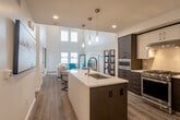 Located at 19550 Vallco Pkwy in Cupertino, CA, this two-story, 2-bedroom, 2-bathroom residence offers a thoughtfully designed layout spread across two floors. The main level features an open-concept living and kitchen area highlighted by soaring double-height ceilings and large clerestory windows that flood the space with natural light and views of the surrounding neighborhood. The kitchen is a standout, equipped with stainless steel appliances including a French door refrigerator, gas range, and built-in microwave, complemented by a spacious center island with a white quartz countertop, undermount sink, and sleek pendant lighting. Dark cabinetry contrasts elegantly with the light hardwood flooring found throughout the main level. The living room benefits from the dramatic ceiling height and ample natural light, creating an open, airy atmosphere. The generously sized primary bedroom features soaring ceilings, mirrored sliding closet doors with built-in shelving and drawers, and hardwood flooring. A second bedroom is bright and comfortable with exterior windows. Both bathrooms are finished with wood-look tile flooring, modern fixtures, and clean white walls, with one featuring a glass-enclosed shower.