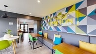 Located at 625 Rue Milton in Montréal, the Terrasse Study Room at Pavillon Milton is a thoughtfully designed communal space that blends energetic aesthetics with functional comfort. The interior is defined by a bold, geometric accent wall in a striking palette of chartreuse, teal, blue, and grey — anchored by an illuminated "Pavillon Milton" sign — creating a dynamic visual centerpiece throughout the space. Large-format light grey tile flooring runs seamlessly across the entire floor, lending a clean, cohesive feel. The main lounge and study area features a mix of seating arrangements, including built-in banquette seating with vibrant accent cushions, sleek wooden tables paired with bright green and yellow modern chairs, and a relaxed lounge zone with upholstered grey armchairs arranged around a round coffee table. A wall-mounted television accompanied by framed artwork and indoor greenery adds warmth to the lounge area. Recessed and pendant lighting provides a well-lit environment suited to both studying and socializing. Generous windows offer panoramic views of the Montréal cityscape and surrounding hillside, flooding the space with natural light. A compact kitchenette with dark cabinetry and a sink provides added convenience. The space also includes a dedicated bicycle storage area, reflecting its practical, resident-focused design.