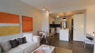 This single-floor unit showcases a thoughtfully designed open-concept layout that integrates the living, dining, and kitchen areas into one cohesive space. The living area is anchored by a clean white sofa, complemented by a woven area rug and a bold orange wall-mounted artwork that adds a vibrant focal point to the neutral palette. Track lighting overhead provides focused illumination throughout the space.

The compact dining area features a round white bistro-style table with wooden chairs, positioned conveniently between the living and kitchen zones. Dark hardwood-style flooring runs consistently throughout, grounding the space with warmth and visual continuity.

The kitchen is well-appointed with white upper cabinetry, stainless steel appliances including a refrigerator, range, and microwave, as well as dark lower cabinets that create a striking contrast. A kitchen island with a light butcher-block-style surface and bar seating offers additional workspace and casual dining. Pendant lighting hangs above the island, enhancing both function and style. The overall ambiance is modern, clean, and efficiently organized.