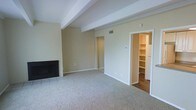 This single-floor space features a generously sized living area finished with neutral-toned carpet that extends throughout the main room, creating a warm and cohesive feel underfoot. A tiled fireplace anchors one wall, serving as a focal point and adding a classic touch to the space. Overhead, an exposed structural beam adds architectural character to the otherwise clean, white ceiling, lending a subtle design element to the room.

The walls are painted in a soft, warm beige tone, contributing to an inviting and neutral ambiance that suits a variety of furnishing styles. A ceiling-mounted HVAC vent indicates a central air system integrated into the layout. Natural light enters through the open areas of the room, keeping the space feeling bright.

Doorways lead to additional areas of the unit, including what appears to be a hallway corridor and a dedicated built-in shelving nook or storage closet outfitted with open wooden shelves and additional cabinetry. The flooring in this adjacent area transitions to hardwood or laminate, offering a practical contrast to the carpeted living space. Overall, the layout is straightforward, functional, and well-suited for comfortable everyday living.