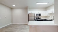 This single-floor unit offers a clean, modern layout designed for comfortable everyday living. The kitchen is a standout feature, outfitted with stainless steel appliances including a refrigerator, range, and over-the-range microwave, complemented by crisp white upper cabinetry, dark lower cabinets, and a light-toned countertop peninsula that provides additional workspace and casual dining potential. Light wood-style luxury vinyl flooring runs throughout the main living and kitchen areas, lending a cohesive, contemporary feel.

The living area is bright and open, anchored by a ceiling fan and a sliding glass door with vertical blinds that invites natural light and provides access to an outdoor space. An adjacent room with matching flooring connects seamlessly to the rest of the unit and offers access to a laundry or utility area.

The bedroom features soft, neutral-toned carpet underfoot, providing warmth and comfort, and includes direct access to an en-suite or nearby bathroom. The bathroom is thoughtfully appointed with a white vanity, integrated sink, a large mirror, and a safety grab bar, reflecting a practical and accessible design. Throughout the unit, neutral warm-beige walls, white trim, and recessed lighting contribute to a clean, inviting atmosphere.