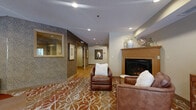This single-floor lobby space is thoughtfully designed to serve as a comfortable and functional common area for residents. The layout flows through several distinct zones, each contributing to an inviting and well-appointed atmosphere. The central lounge area anchors the space with a wood-mantled fireplace flanked by leather armchairs arranged atop a richly patterned terracotta and ivory area rug, creating a warm and welcoming focal point. Recessed lighting throughout provides consistent illumination, complementing the neutral beige and taupe wall tones.

Adjacent to the lounge, a dedicated mail center features a built-in bank of individual mailboxes framed beneath a decorative "MAIL" sign, accompanied by a wood-shelf counter and hardwood flooring that extends through the transitional corridors. The elevator lobby is accessed through a textured-wall corridor adorned with framed artwork and a cushioned bench for convenience.

A separate sunlit seating alcove offers two upholstered chairs positioned beside large windows with white blinds, allowing natural light to fill the space and provide views of outdoor greenery. Lush potted plants enhance the organic, residential feel. Patterned carpet ties the lounge and seating zones together, while smooth hardwood floors define the more functional circulation areas.
