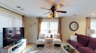 This 2-floor home offers a thoughtfully designed living space that balances comfort and style. The living room serves as the heart of the main level, showcasing warm beige walls accented by crown molding that lends a classic, polished character to the space. A decorative five-blade ceiling fan with amber glass light fixtures serves as a functional centerpiece, providing both lighting and airflow.

Natural light flows generously into the room through multiple windows dressed with neutral linen drapes and white plantation-style shutters, creating a bright and airy atmosphere. The flooring features light hardwood that complements the room's warm color palette.

Seating is arranged thoughtfully, with a pair of white tufted accent chairs flanking a small wooden side table, positioned near the windows to take advantage of the natural light. A plush patterned area rug in neutral tones anchors the seating arrangement, adding texture and warmth underfoot. A rich purple sofa adds a bold contrast to the otherwise neutral palette. Wall décor, including a sunburst mirror and arrow art, contributes to the space's casual yet curated aesthetic.