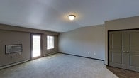 This single-floor space features a spacious, open living area designed with simplicity and functionality in mind. The room is finished with wall-to-wall plush carpeting in a neutral tone, providing a comfortable and cohesive foundation throughout. Walls are painted in a soft greige hue, creating a warm yet understated backdrop that complements a wide range of décor styles.

Natural light is a notable feature of this space, provided by a large sliding glass patio door paired with an adjacent window along one wall. Together, these openings allow ample daylight to fill the room and offer views of the outdoors, contributing to an airy and open ambiance. The patio door also provides convenient exterior access, adding to the overall functionality of the space.

A baseboard heating unit runs beneath the windows, ensuring efficient climate control throughout the room. Overhead, a flush-mount ceiling light fixture provides general illumination, complemented by the natural light during daytime hours. Dark baseboards contrast neatly against the lighter walls and carpet, adding a subtle architectural definition. The overall layout is clean, open, and well-suited for comfortable everyday living.