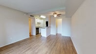 This single-story space features an open-concept layout that connects the living area to the kitchen, creating a cohesive and functional flow throughout. The floors are finished with warm-toned hardwood-style planking that extends seamlessly across the main living and kitchen areas, contributing to a clean, consistent aesthetic.

The living area is well-proportioned and benefits from a ceiling fan with an integrated light fixture, providing both comfort and ambient lighting. Neutral white walls and a textured ceiling give the space a bright, airy feel.

The kitchen is positioned at the rear of the main living space and is partially separated by a peninsula-style counter, offering both a visual divider and additional workspace. White cabinetry lines the upper walls, complemented by stainless steel appliances including a dishwasher and refrigerator. A utility sink and countertop workspace complete the kitchen's practical layout.

Multiple doorways visible from the main living area suggest access to additional rooms, including what appears to be a hallway leading to further private spaces. Overall, the unit presents a functional, well-maintained interior with a modest, clean design.